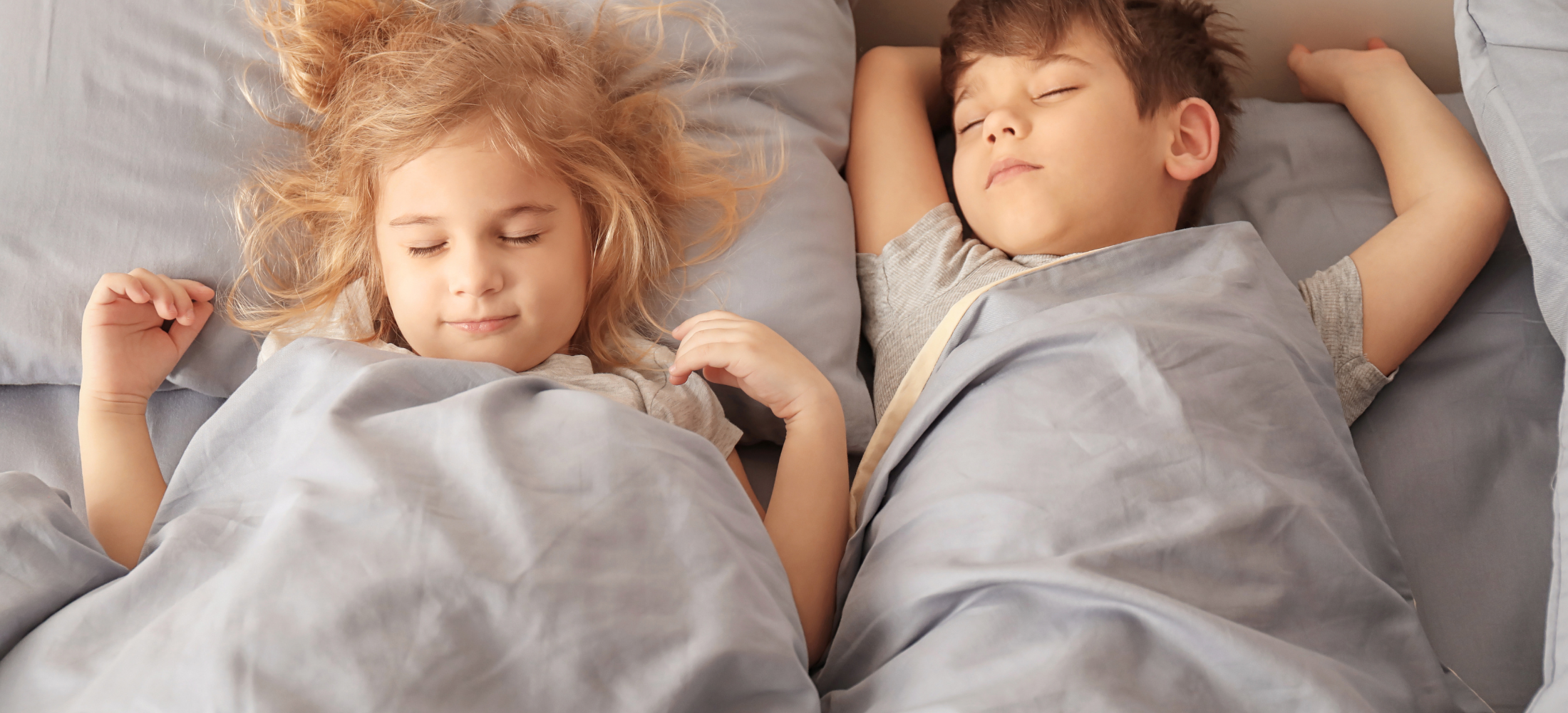Two children sleeping peacefully under grey bedding.