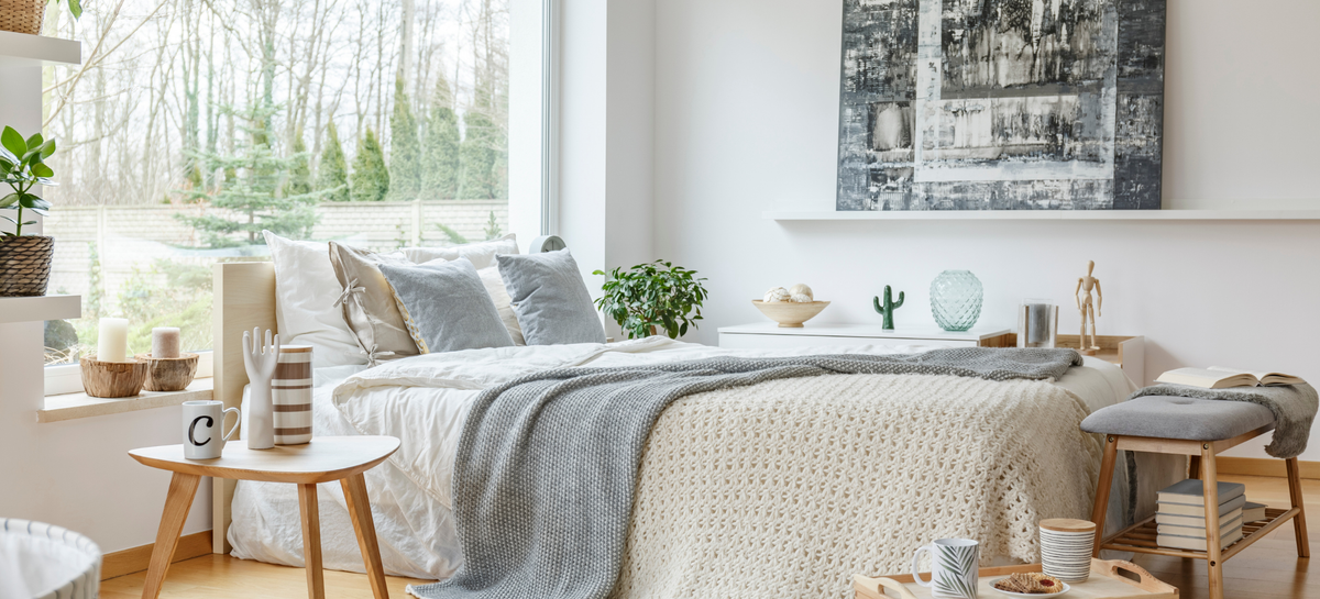 Bright bedroom with Yorklinen microfiber duvet by Maholi Inc bedding, layered neutral pillows and grey and cream knit blankets beside a large window overlooking trees.
