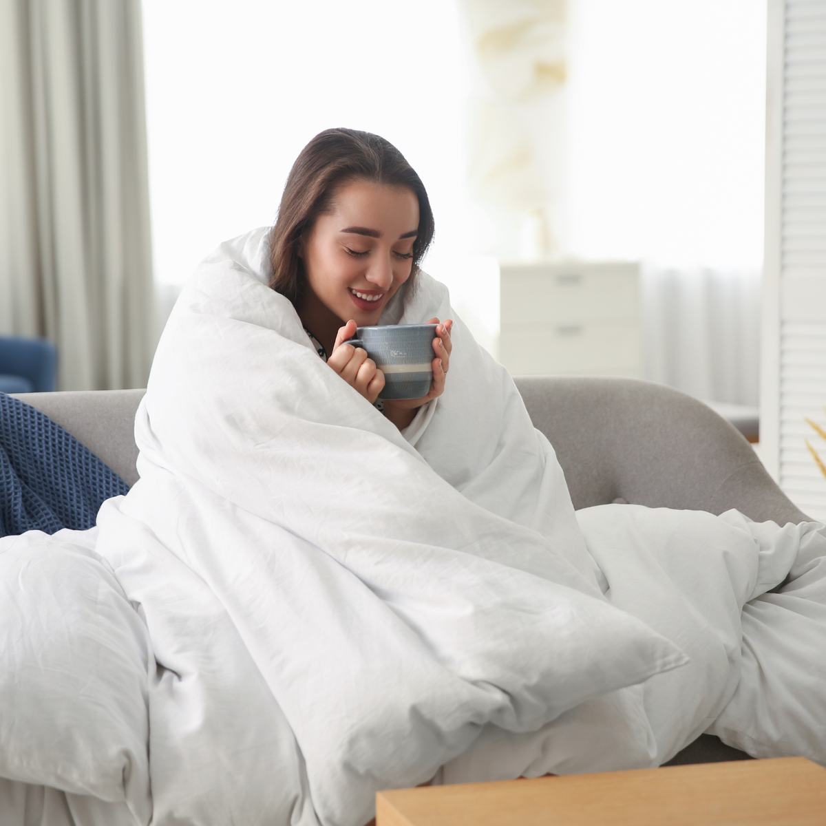 Woman wrapping the best Royal Elite white goose down duvet feeling cozy and holding coffee mug.