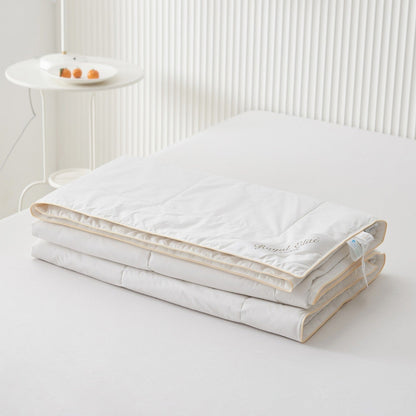Royal Elite Silk Duvet folded on a bed, showing its low profile, gold piped edge and embroidered logo.